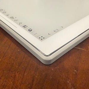 May include: A white digital tablet with a gray frame, viewed at an angle. The screen is bright and clean, with icons and navigation controls visible. The device rests on a wooden surface, highlighting its modern aesthetic.
