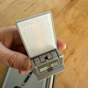 Può includere: Un accessorio in plastica grigio chiaro con un pannello bianco rettangolare testurizzato, una base incernierata, due punti di contatto metallici rettangolari e tre piccoli pin color oro. L'accessorio presenta il testo in rilievo "SUPER NOTE".