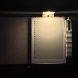 May include: A white e-reader with a stylus and a cover, lit by a small lamp. The e-reader's screen shows a menu of icons. The stylus is white and has a textured design. The cover is a dark grey colour.