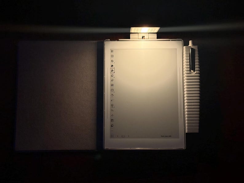 May include: A white e-reader with a stylus and a cover, lit by a small lamp. The e-reader's screen shows a menu of icons. The stylus is white and has a textured design. The cover is a dark grey colour.