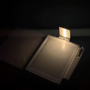 May include: An illuminated e-reader with an integrated light and a protective cover, open and angled. The screen is visible, and the background is dark. A focused light source highlights the e-reader.