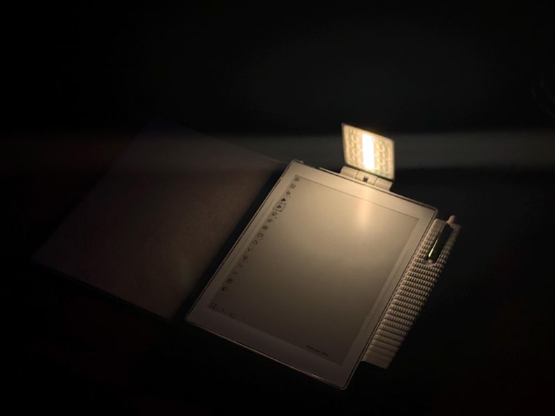 May include: An illuminated e-reader with an integrated light and a protective cover, open and angled. The screen is visible, and the background is dark. A focused light source highlights the e-reader.