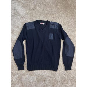 May include: A black V-neck jumper featuring reinforced fabric patches on the shoulders and elbows, along with a small rectangular chest pocket. The garment includes a ribbed collar, cuffs, and hem, with a small white brand label at the neckline.