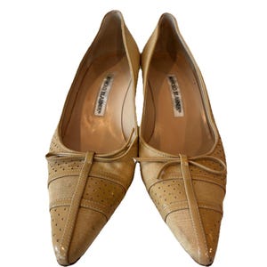 Puede incluir: Un par de zapatos de tacón beige Manolo Blahnik con punta. Los zapatos presentan un detalle de lazo, detalles perforados y una tela texturizada. Los tacones son bajos y los zapatos están sobre un fondo blanco.