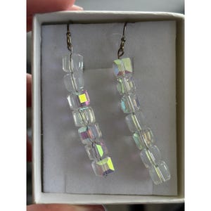 May include: A pair of clear, iridescent cube earrings. Each earring has a series of clear, square beads strung together, with a subtle rainbow sheen. The earrings are displayed in a white box, showcasing their delicate design.