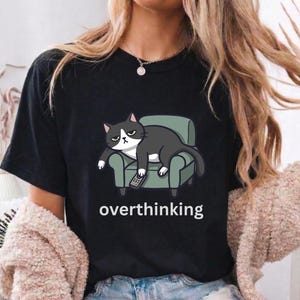 Puede incluir: Camiseta negra con un gato de dibujos animados en un sillón verde, sosteniendo un mando a distancia. El gato tiene una expresión de mal humor. La palabra "overthinking" está impresa en blanco debajo de la ilustración del gato. La camiseta la lleva una persona.