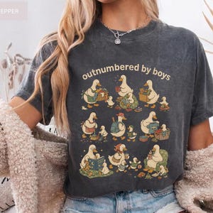 May include: Charcoal grey t-shirt with a cartoon duck family design and the words "outnumbered by boys". The ducks are shown in different scenes, including reading and carrying baskets. The t-shirt has a faded, vintage appearance.
