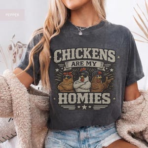 May include: Dark gray t-shirt with the text "CHICKENS ARE MY HOMIES." The graphic features three cartoon chickens wearing sunglasses and hats. The shirt is a casual, unisex style.