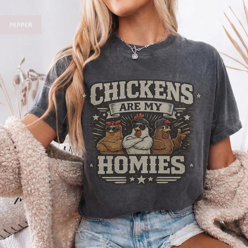May include: Dark gray t-shirt with the text "CHICKENS ARE MY HOMIES." The graphic features three cartoon chickens wearing sunglasses and hats. The shirt is a casual, unisex style.