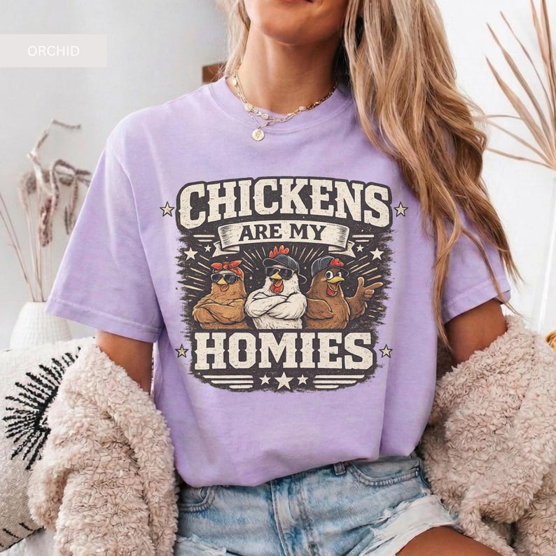 May include: A light purple t-shirt with the text "CHICKENS ARE MY HOMIES" in a distressed font. The graphic features three cartoon chickens wearing sunglasses and bandanas. The shirt is a casual wear item.