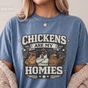 May include: Blue jean-colored t-shirt with the text "CHICKENS ARE MY HOMIES" in a distressed font. The graphic features three chickens wearing sunglasses and bandanas. A fun, casual tee.