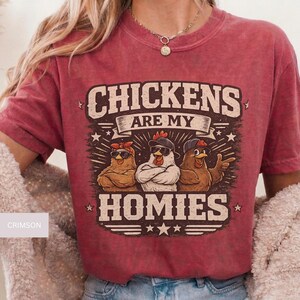 May include: Crimson-colored t-shirt featuring a graphic of three chickens wearing sunglasses and hats. The text "CHICKENS ARE MY HOMIES" is displayed above the chickens. The shirt has short sleeves and a crew neck.