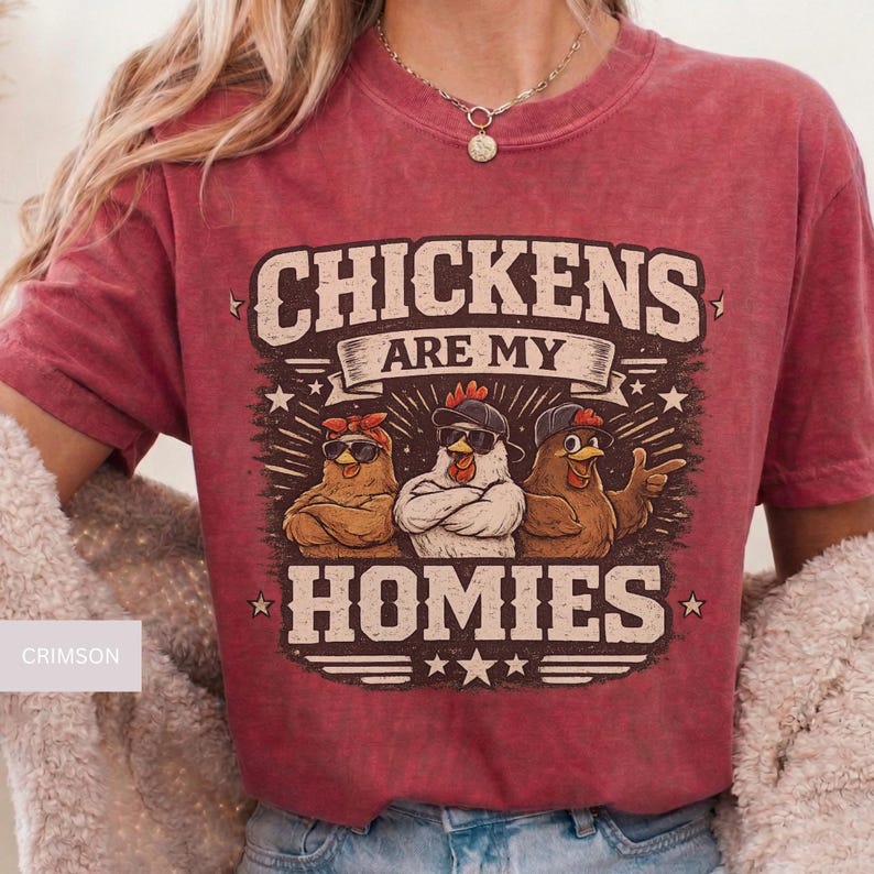 May include: Crimson-colored t-shirt featuring a graphic of three chickens wearing sunglasses and hats. The text "CHICKENS ARE MY HOMIES" is displayed above the chickens. The shirt has short sleeves and a crew neck.