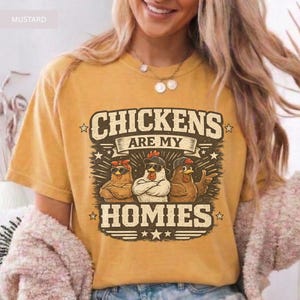 May include: Mustard-colored t-shirt with the text "CHICKENS ARE MY HOMIES" in a distressed font. The graphic features three chickens wearing sunglasses and hats. The shirt is a short-sleeved crew neck.