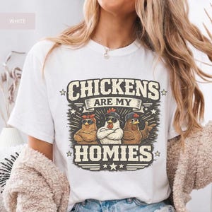 May include: White t-shirt with the text "CHICKENS ARE MY HOMIES" in a distressed, vintage style. The graphic features three cartoon chickens wearing sunglasses and bandanas. The design is surrounded by stars and decorative elements.
