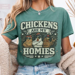 May include: Light green t-shirt with the text "CHICKENS ARE MY HOMIES" above a graphic of three chickens wearing sunglasses and hats. The shirt is a solid color and has a vintage, worn look.