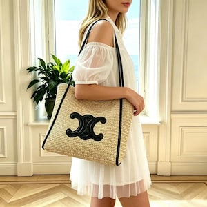May include: A beige woven tote bag with black leather trim and handles. The bag features a large black emblem on the front. The bag is rectangular and appears to be carried over the shoulder.