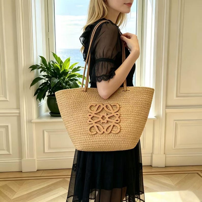 May include: A woven straw tote bag with leather handles and a decorative emblem. The bag is a light tan color and appears to be spacious. The bag is held by a person wearing a black dress.
