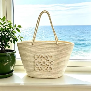 May include: A woven beige tote bag with cream-colored handles and a decorative emblem. The bag is set against a backdrop of a window overlooking the ocean and a potted plant.