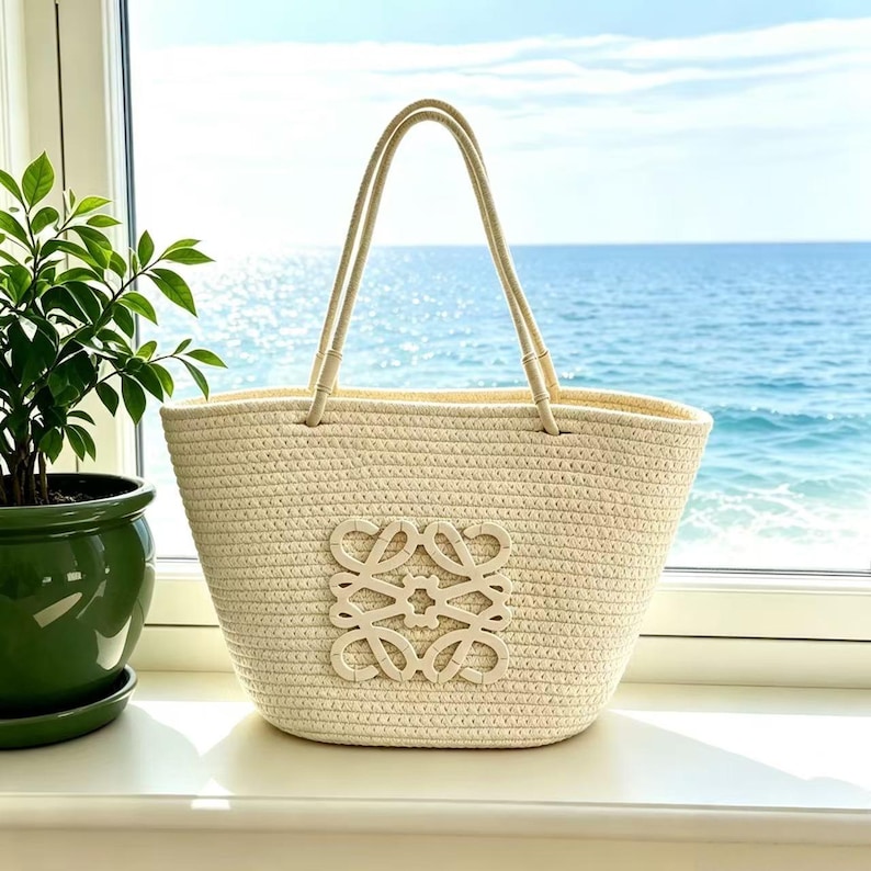 May include: A woven beige tote bag with cream-colored handles and a decorative emblem. The bag is set against a backdrop of a window overlooking the ocean and a potted plant.