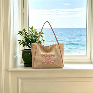 May include: A beige woven tote bag with pink leather trim and handles. The bag features a pink logo on the front. The bag is on a white windowsill with a view of the sea and a green potted plant.