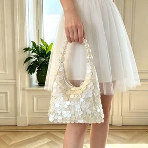 May include: A white, sequined handbag with a pearl handle is held by a person. The bag is covered in round, iridescent sequins. The handle is made of large, round pearls. The person is wearing a white dress.