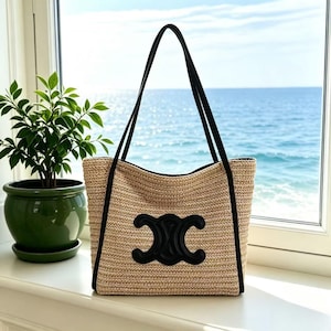 May include: A beige woven tote bag with black trim and handles. The bag features a large black emblem on the front. The background shows a window with a view of the sea and a potted plant.