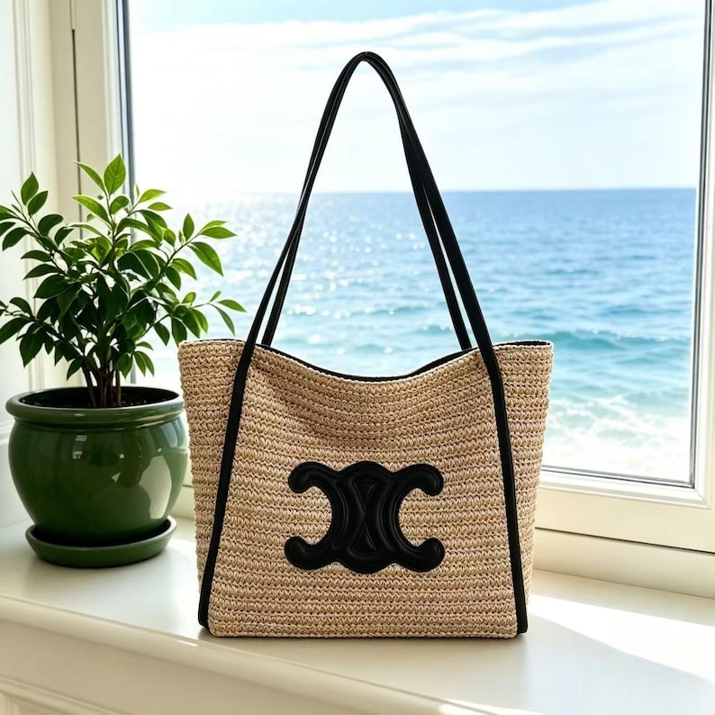 May include: A beige woven tote bag with black trim and handles. The bag features a large black emblem on the front. The background shows a window with a view of the sea and a potted plant.