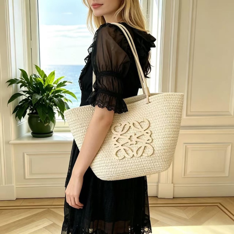 May include: A cream-colored woven tote bag with a large, light-colored logo on the front. The bag has cream-colored handles and is rectangular in shape. The bag is made of a woven material.