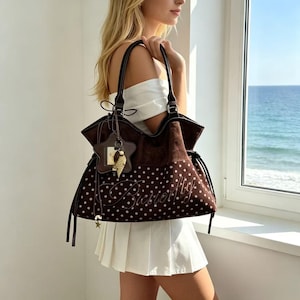 K&ouml;nnte beinhalten: Eine braune Tragetasche mit wei&szlig;en Punkten und dem Wort "Butterfly" auf der Vorderseite gestickt. Die Tasche hat dunkelbraune Griffe und einen sternf&ouml;rmigen Anh&auml;nger. Die Tasche wird von einer Person getragen.