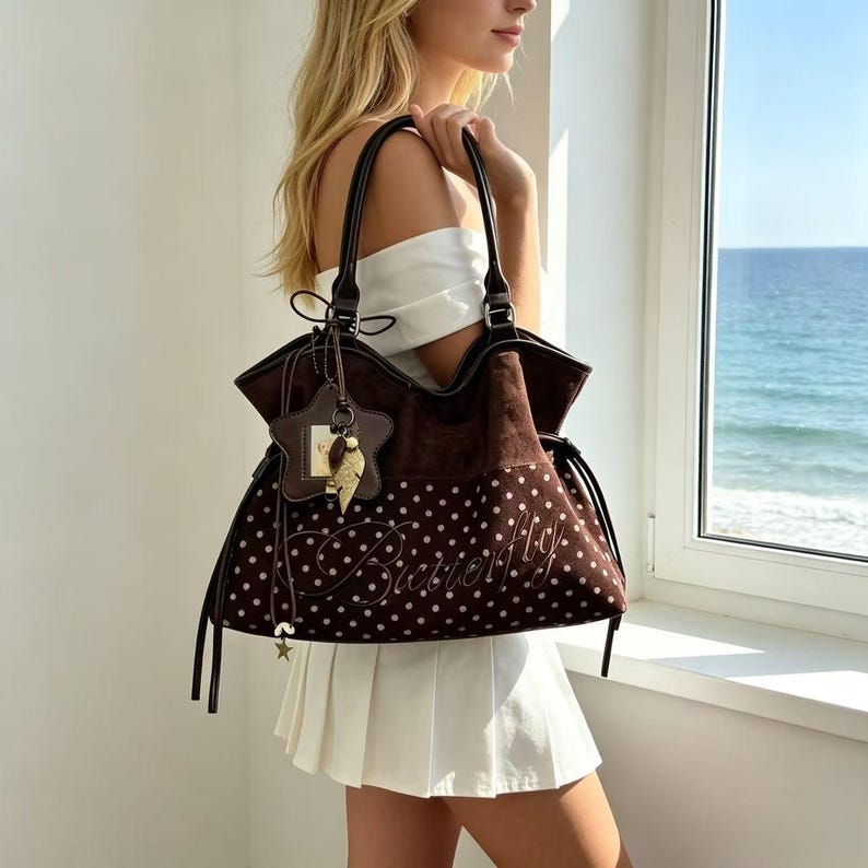 K&ouml;nnte beinhalten: Eine braune Tragetasche mit wei&szlig;en Punkten und dem Wort "Butterfly" auf der Vorderseite gestickt. Die Tasche hat dunkelbraune Griffe und einen sternf&ouml;rmigen Anh&auml;nger. Die Tasche wird von einer Person getragen.
