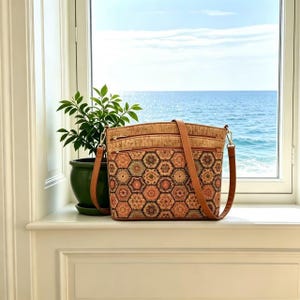 May include: A cork crossbody bag with a geometric pattern of hexagons in shades of brown, orange, and green. The bag has a brown leather strap and zipper closures. The bag is sitting on a white windowsill with a view of the ocean.