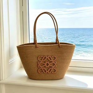 May include: A woven straw tote bag with brown leather handles and a brown leather emblem. The bag is tan and sits on a white windowsill with a view of the ocean.