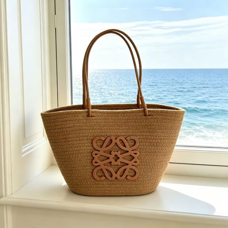 May include: A woven straw tote bag with brown leather handles and a brown leather emblem. The bag is tan and sits on a white windowsill with a view of the ocean.