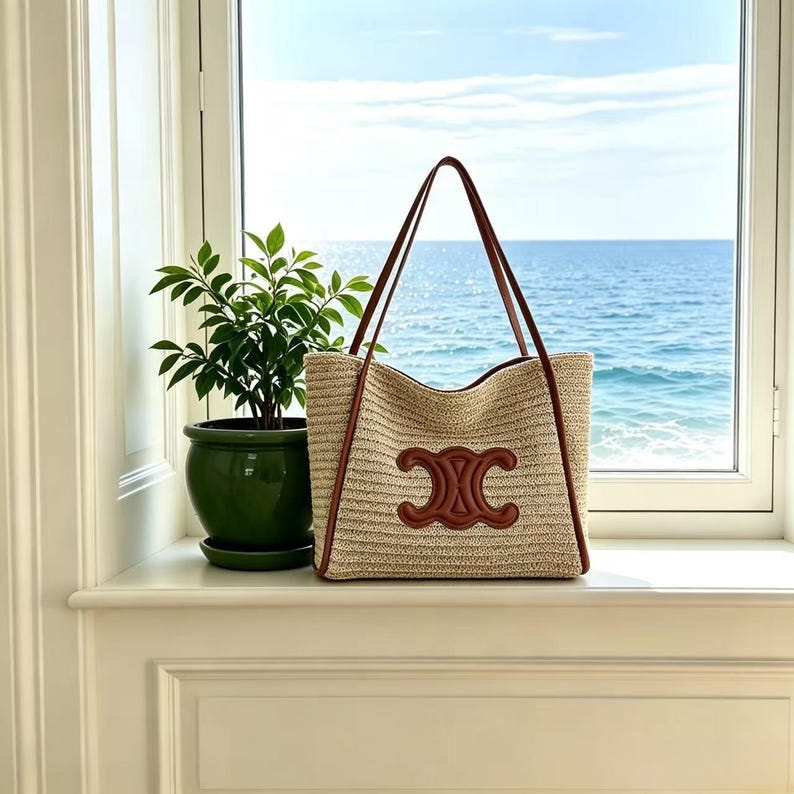 May include: A beige woven tote bag with brown leather handles and trim. The bag features a large brown emblem on the front. The bag is sitting on a white windowsill with a view of the sea and a green potted plant.