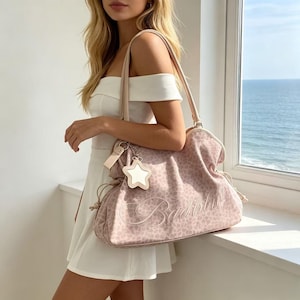 K&ouml;nnte beinhalten: Eine hellrosa Handtasche mit Leopardenmuster und dem Wort "Babydoll" auf der Vorderseite gestickt. Die Tasche hat einen sternf&ouml;rmigen Anh&auml;nger und einen hellrosa Riemen. Die Tasche wird von einer Person gehalten, die ein wei&szlig;es Kleid tr&auml;gt.