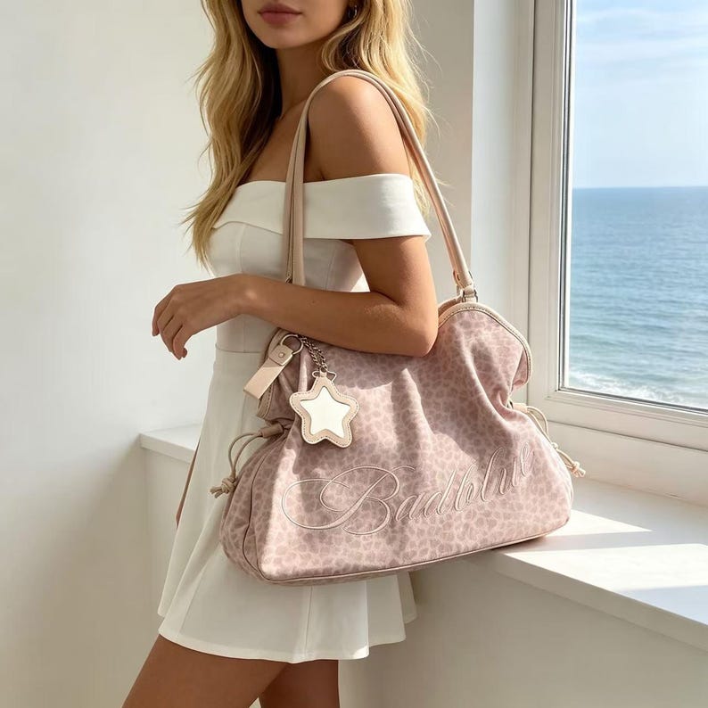 K&ouml;nnte beinhalten: Eine hellrosa Handtasche mit Leopardenmuster und dem Wort "Babydoll" auf der Vorderseite gestickt. Die Tasche hat einen sternf&ouml;rmigen Anh&auml;nger und einen hellrosa Riemen. Die Tasche wird von einer Person gehalten, die ein wei&szlig;es Kleid tr&auml;gt.