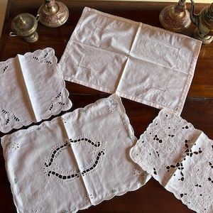 May include: A collection of white, intricately embroidered linens, including placemats and doilies, are arranged on a dark wooden surface. The linens feature various cut-out and floral designs, showcasing a vintage aesthetic. Decorative metal objects are in the background.