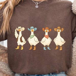 May include: A brown t-shirt with a cartoon design of four geese in cowboy attire. Each goose is depicted wearing a cowboy hat, boots, and a bandana. The bandanas are in different colours. The design is centered on the shirt.