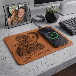 May include: A rectangular tan leather wireless charging mouse pad featuring a laser-engraved portrait of a person holding a dog, with the text "Best Dad Ever!" written in cursive script. A black smartphone is charging on the right side of the pad.