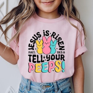 May include: A pink t-shirt with short sleeves, displaying the words "JESUS IS RISEN" above three cartoon Easter bunnies in yellow, pink, and blue. The text "TELL YOUR PEEPS!" is below in colourful letters.