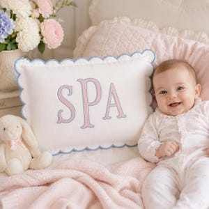 May include: A rectangular white decorative pillow with a scalloped blue edge and the letters SPA embroidered in pink with blue outlines. The pillow is positioned next to a plush cream-colored rabbit toy and a baby wearing a white floral onesie.