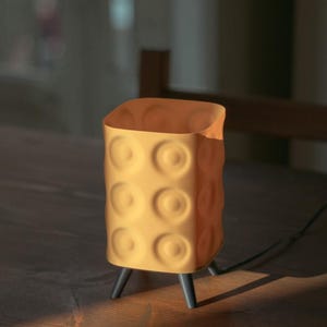May include: A yellow table lamp with a textured surface and a square shape. The lamp has a series of circular indentations and stands on three black legs. A black cord extends from the base.