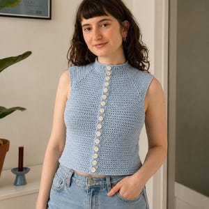 Puede incluir: Un top de ganchillo sin mangas en color azul claro con cuello alto y una hilera vertical de pequeños botones redondos de color claro en la parte delantera. La prenda está confeccionada con un patrón de punto uniforme y corte ajustado.