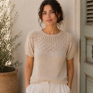 May include: A beige short-sleeved knit top featuring a round neckline with ribbed detailing and a delicate diamond-shaped openwork pattern across the yoke and shoulders. The garment has a smooth, solid-knit body and a relaxed, comfortable fit.