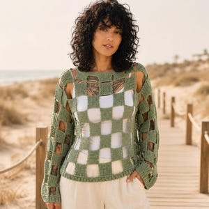 May include: A sage green long-sleeve crochet sweater featuring a square checkerboard pattern with openwork cutouts throughout the body and sleeves. The garment has a round neckline and is worn over a plain white base layer with light-colored trousers.