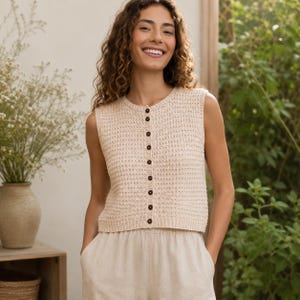 Op de afbeelding: Een mouwloos gebreid vest in crèmekleur met een ronde halslijn en een knoopsluiting aan de voorzijde met kleine, donkerbruine ronde knopen. Het kledingstuk heeft een gestructureerd breipatroon en een rechte zoom die op de taille valt.