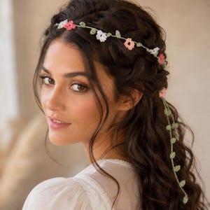 Puede incluir: Un delicado accesorio para el cabello tejido a crochet con una fina banda verde similar a una enredadera, adornada con pequeñas flores multicolores en tonos rosa, blanco y melocotón. Incluye detalles de hojas verdes que cuelgan a un lado.