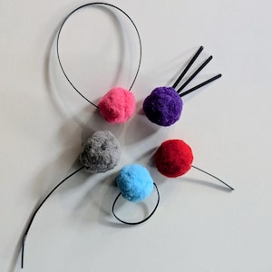 May include: A collection of colorful pom-pom hair accessories. The image shows a variety of pom-poms in pink, purple, gray, red, and blue, each attached to a black headband or stem. The background is a plain white surface.