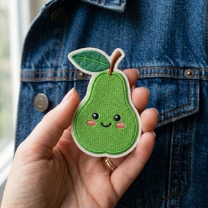 May include: A green pear-shaped embroidered patch with a smiling face, blush cheeks, and a brown stem and leaf. The patch is held in front of a blue denim jacket.